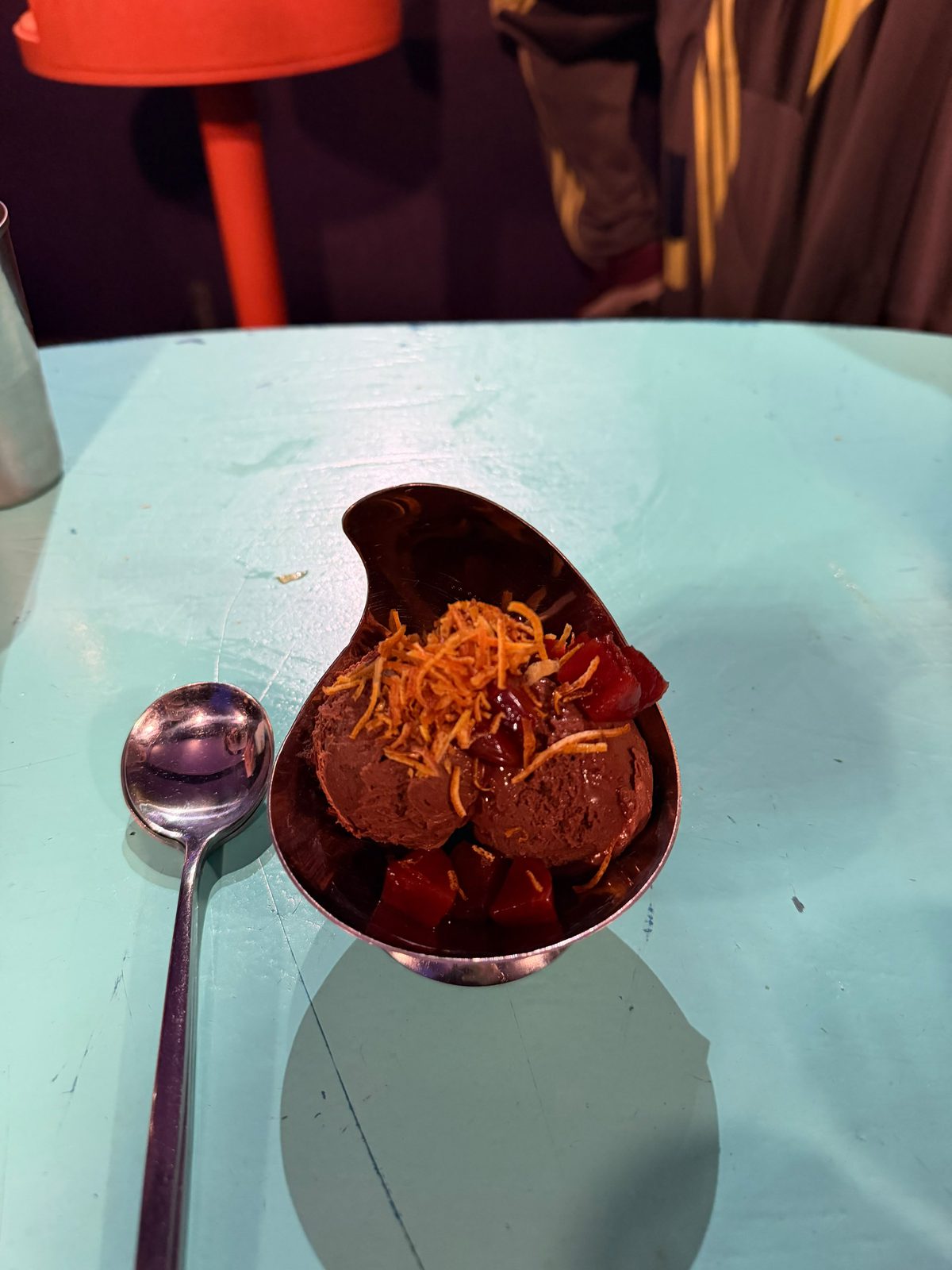 Mousse de chocolate de Cang Tin en una copa de metal, con hebras crocantes por encima, sobre una mesa turquesa
