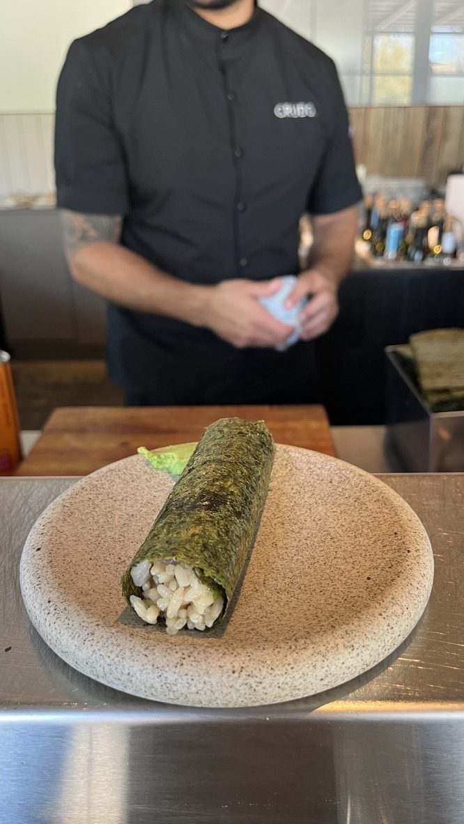 Handroll de pesca blanca con furikake ahumado, tajín y negi sobre un plato de piedra, con wasabi al costado, en la barra de Crudo Barra