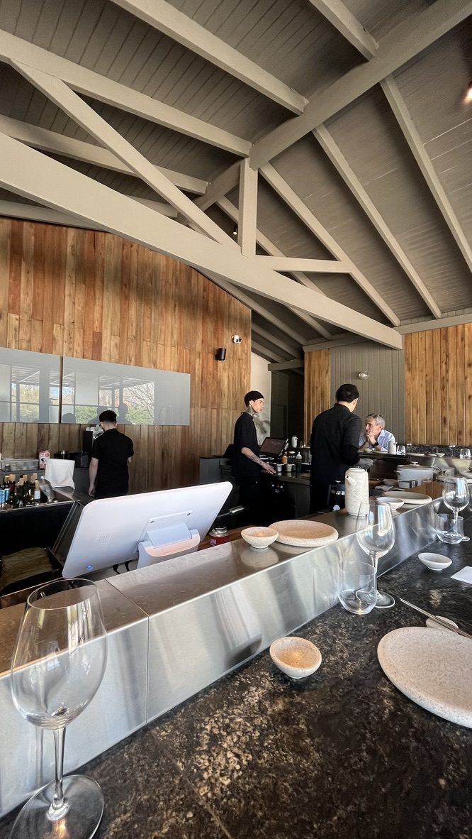 La barra de acero de Crudo Barra en La Aldea, Pilar, con las paredes de madera, el techo de vigas blancas y los cocineros trabajando detrás