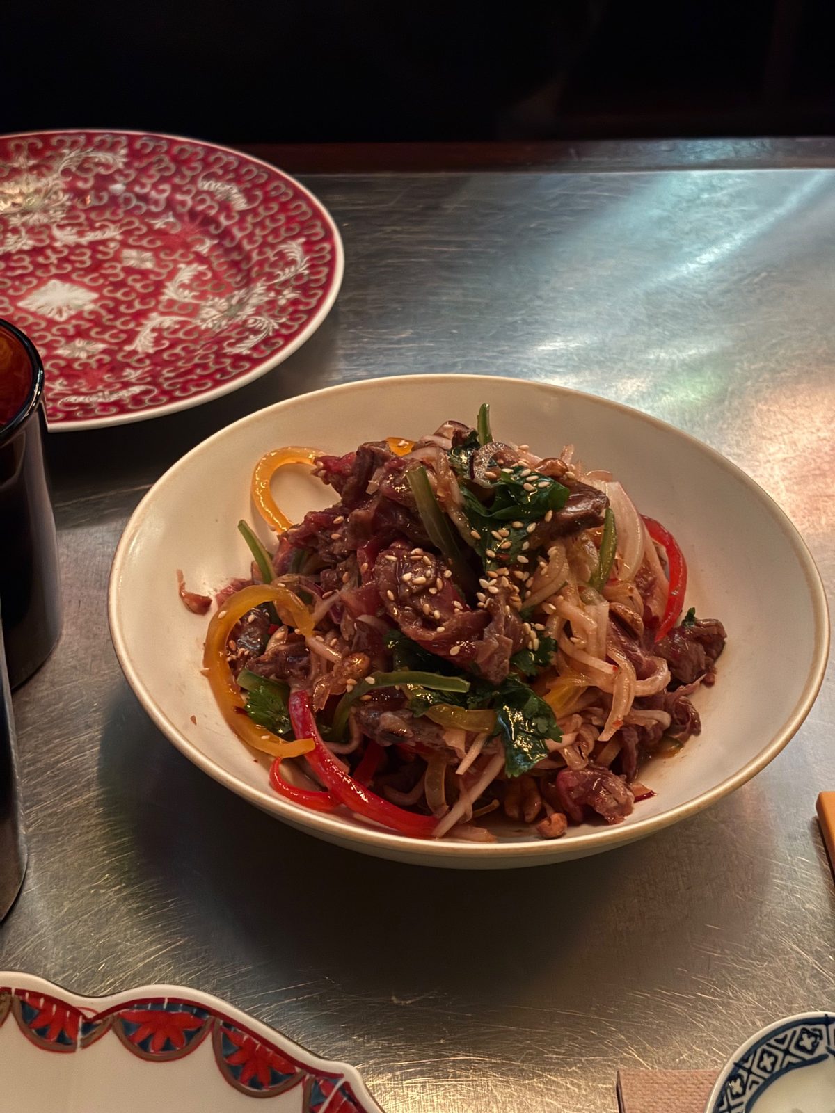 Ensalada de carne de Tony Wu en un bol blanco: tiras de carne con morrones, cebolla, cilantro, maní y sésamo, sobre una mesa de acero